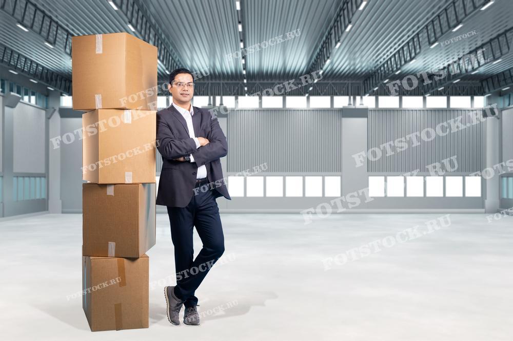 На изображении показан мужчина, стоящий рядом с несколькими сложенными коробками в современном складском помещении. Он готовит свои личные вещи к временному хранению в индивидуальной ячейке. Современные складские условия обеспечивают удобство и безопасность для хранения.